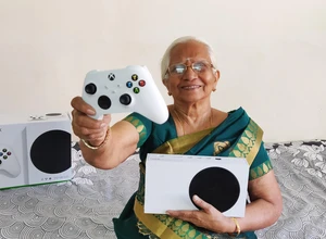 Пожилая женщина в сари держит игровой контроллер и консоль Xbox на диване.