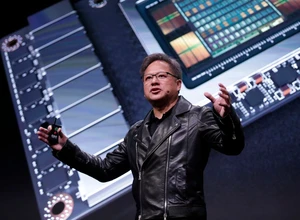 Мужчина в черной куртке выступает на сцене с технологической презентацией за ним.