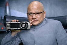 Мужчина в сером костюме держит видеокарту, задумчиво прикасаясь ко рту.